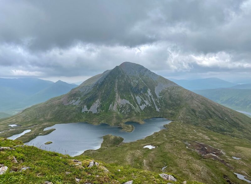 The whole hill, a lone dark cone with pale scree patches, above a grey lochan under heavy cloud, seen from a grassy slope with stones in the foreground.