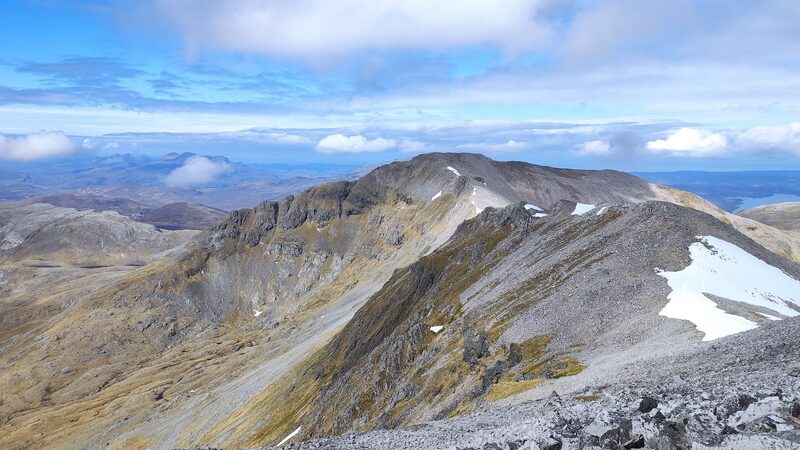 The grey stony ridge between Ben More Assynt and Conival running to a rounded summit, crags and snow patches on its flank and hills far beyond.