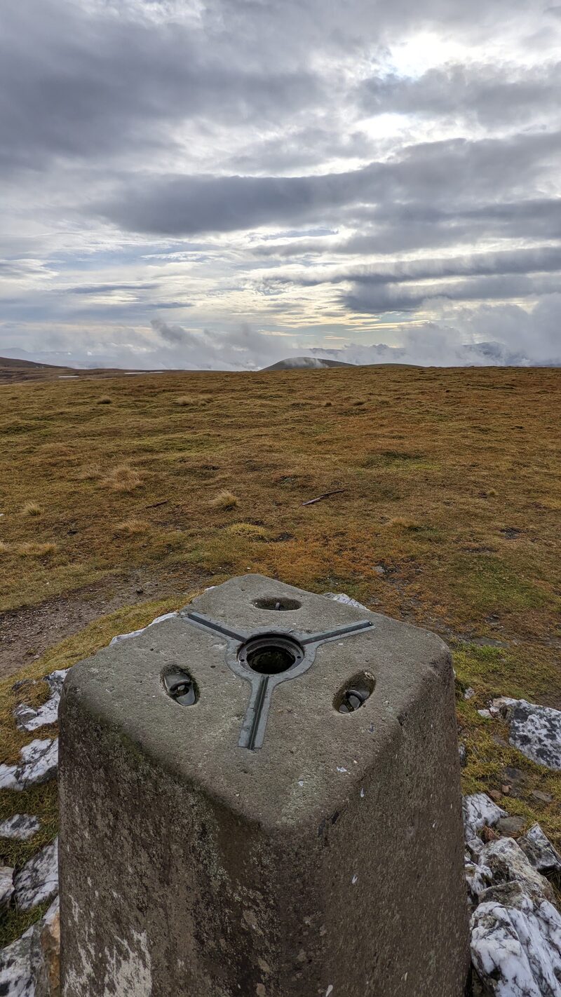 The top of the trig pillar close to, with the wide grassy plateau beyond and cloud lying on far hills.