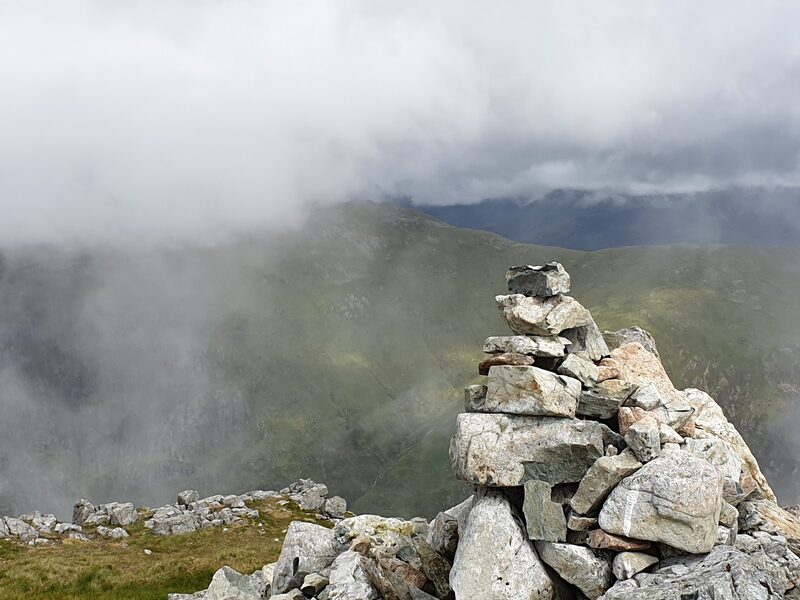 A summit cairn of piled stones, with cloud blowing across the hillside behind.