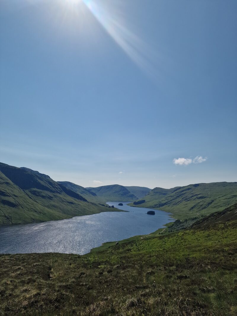 A long loch with two small islands between steep green hillsides, seen from the slope above it.