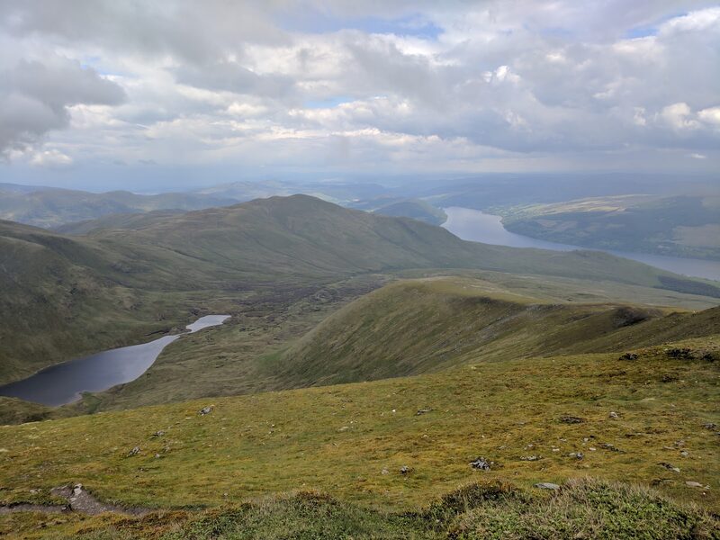 A rounded green hill above a lochan, seen from higher ground, with a long loch beyond under broken cloud.