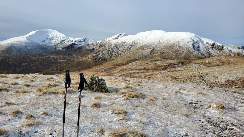 A small cairn on a rounded, frosted top with two walking poles stood beside it, and three snow-covered hills beyond under grey cloud.