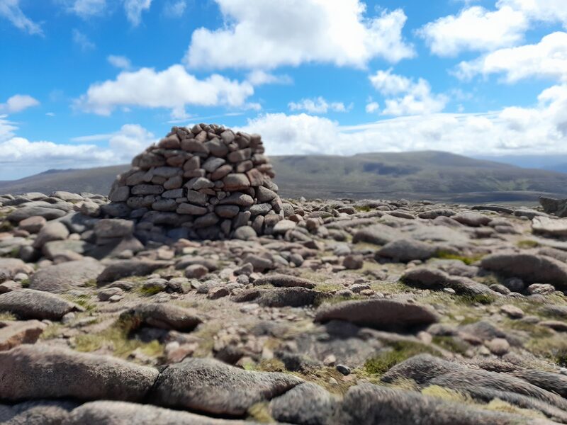 The tall beehive summit cairn of pink granite on a top of pink boulders, a long rounded hill behind under blue sky and white cloud.