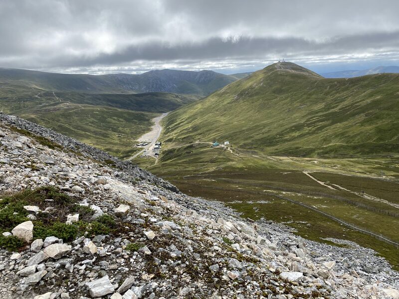 The view from Càrn Aosda's pale scree down to the Glenshee ski centre and the A93, with The Cairnwell and its masts across the pass under heavy cloud.