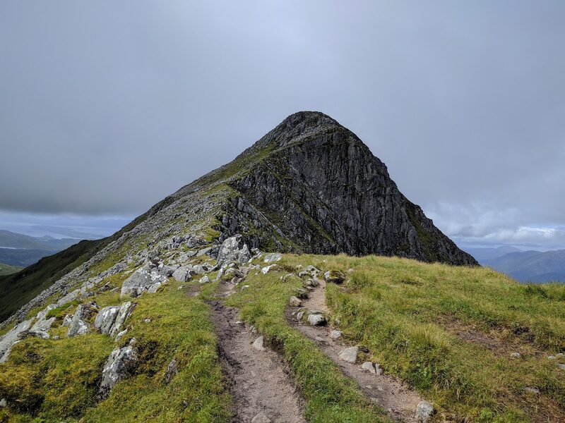 Sgorr Dhonuill seen along the ridge: a steep peak with a wall of dark crag on one side, a worn path running towards it over grass.