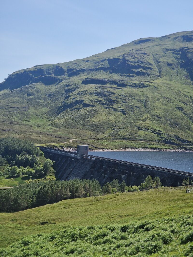 A concrete dam holding back a loch beneath a steep green hillside banded with crags.