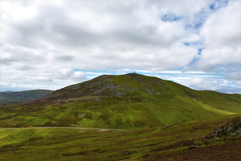 The Cairnwell, a green and stony hill with its masts on top, above a track across the moor under broken cloud.