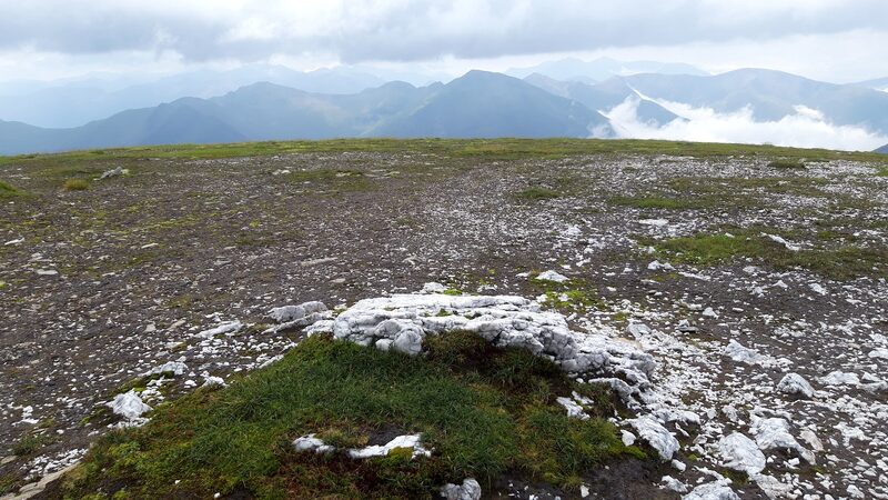 A broad, stony, mossy top with pale rocks in the foreground and a line of peaks beyond under cloud.