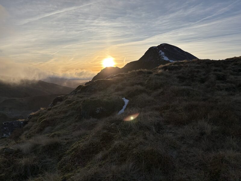 The peak in silhouette with the sun rising beside it, seen from a grassy shoulder.