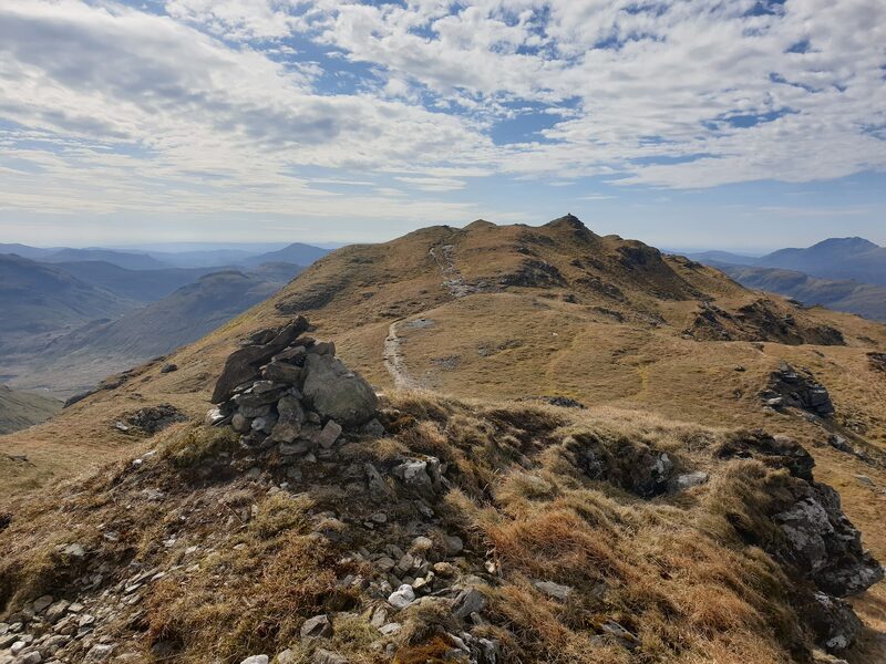 A small cairn on a knoll of Beinn Chabhair's summit ridge, a worn path running on along the rocky crest to a higher top under a dappled sky.