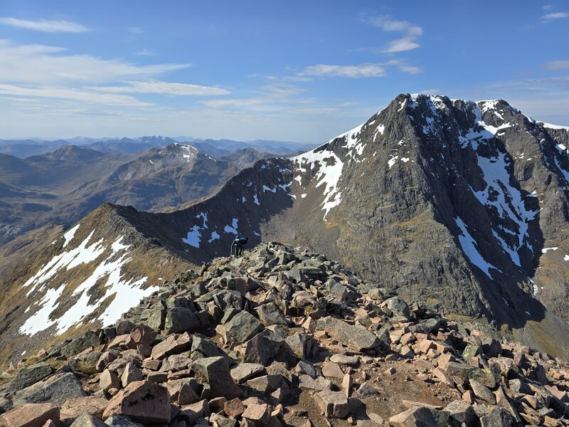 The summit and its cliffs with snow patches, seen along a rocky ridge, with one distant figure.