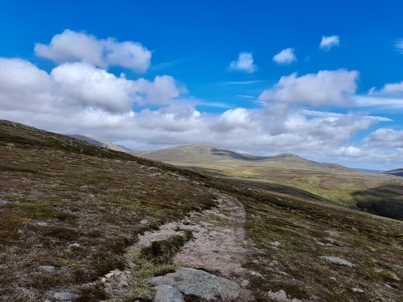 A stony path over a heather shoulder with a rounded hill ahead.