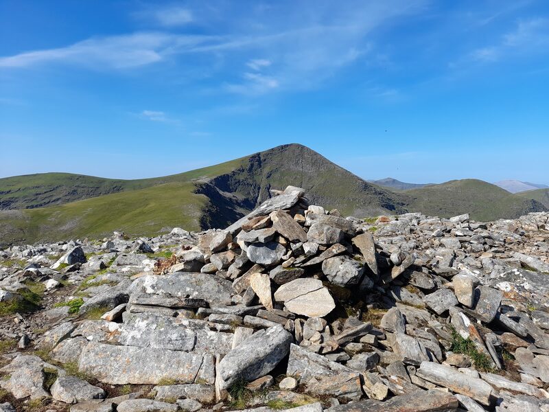 Sgurr Mor's sharp cone, dark crags on one flank, standing above the rest of the range under a blue sky, seen across a low pile of stones on a neighbouring top.