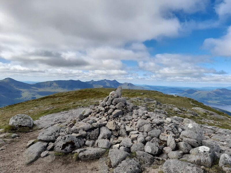 The summit cairn of granite blocks with a pointed stone on top, a jagged ridge on the skyline to the left and a sea loch far below to the right.