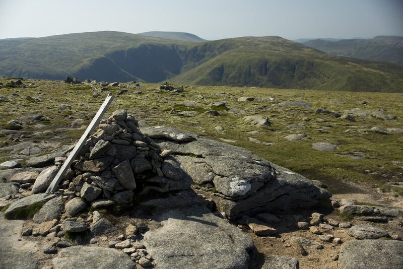 A cairn built on a granite slab on the stony top of Carn an t-Sagairt Mor, a strip of metal leaning against it, with a deep glen and rounded hills beyond.