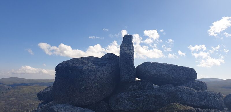Big granite blocks stacked on the top of Broad Cairn against a blue sky with white cloud, moor far below.