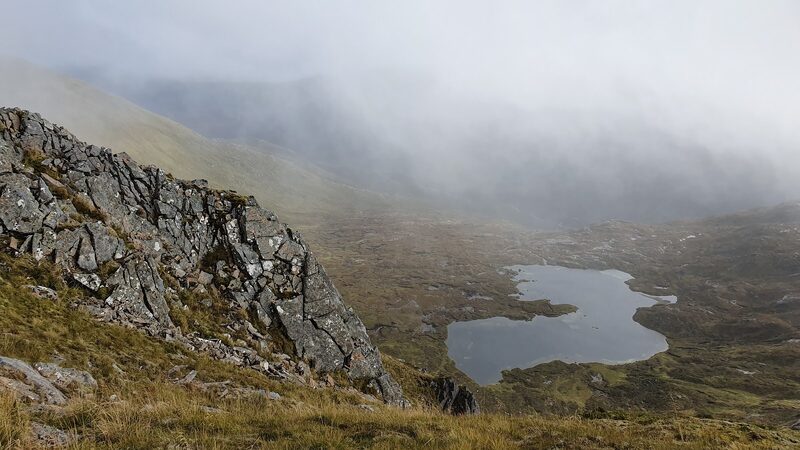 A dark crag above a lochan on a corrie floor, with mist drifting over the slopes behind.