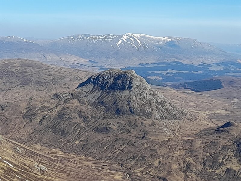 The view from high on the hill down to a craggy knoll standing alone in a wide brown glen, a long snow-streaked range beyond under a clear sky.