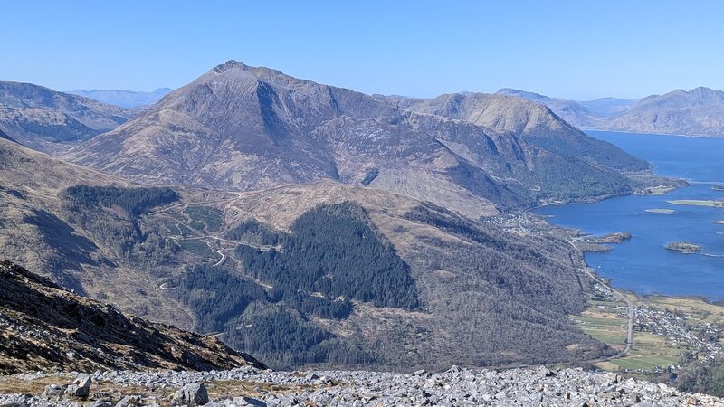 Beinn a' Bheithir seen from the east: its pointed eastern peak above forest, with Ballachulish on the shore of Loch Leven below and the loch running out to the bridge.