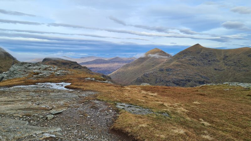 Two neighbouring summits seen across a rocky top with a small pool, a far range blue beneath them.