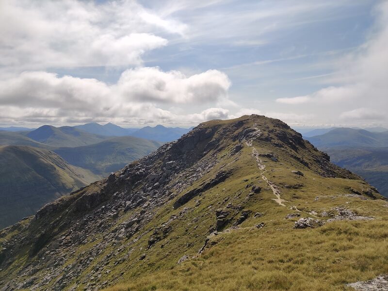 A path running along a grassy, rocky ridge to a summit, with blue hills beyond under broken cloud.