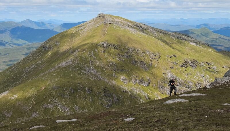 A walker crossing a grassy shoulder below a steep green cone, with blue ranges beyond.