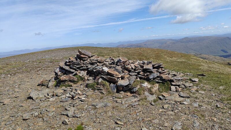 The summit cairn of Beinn Sheasgarnaich, a low wide ring of flat grey and rust-coloured slabs on a bare stony top, with a long line of hills far off under a blue sky.