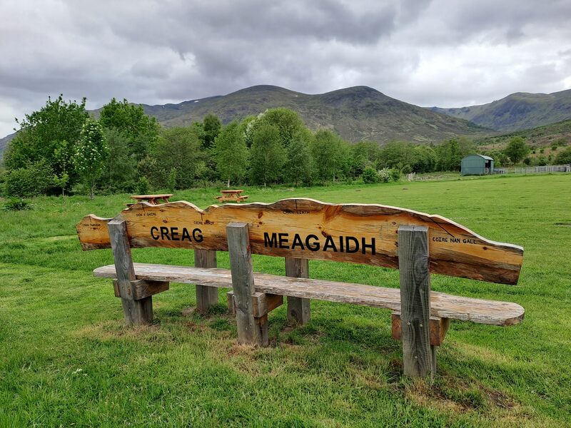 A rough-sawn wooden bench with CREAG MEAGAIDH carved along its back, on the grass at Aberarder, wooded slopes and rounded hills under heavy cloud behind.
