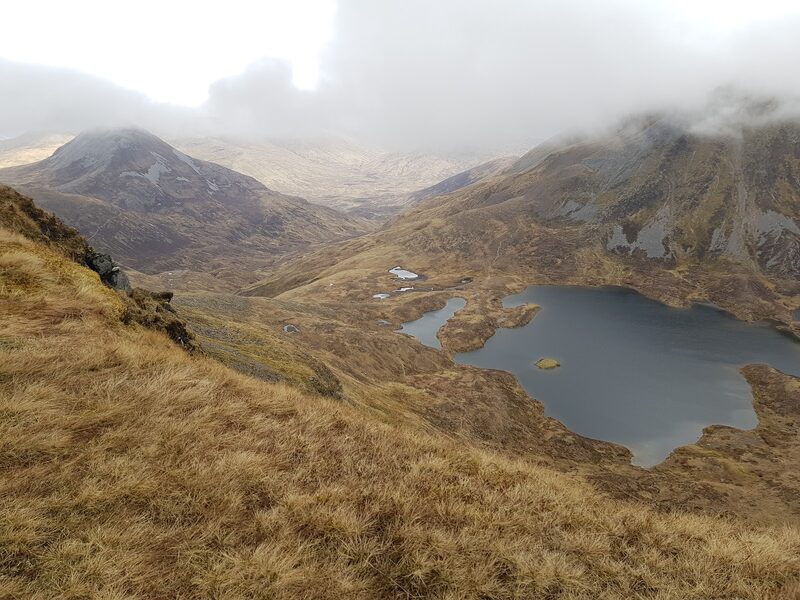 From a tawny slope of the hill, a dark lochan in a high hollow and a pale, cloud-capped hill beyond.