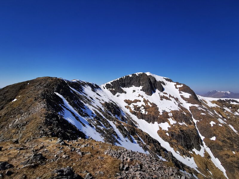 The summit and the dark crag under it, streaked with spring snow, seen along the ridge from a stony shoulder under a deep blue sky.