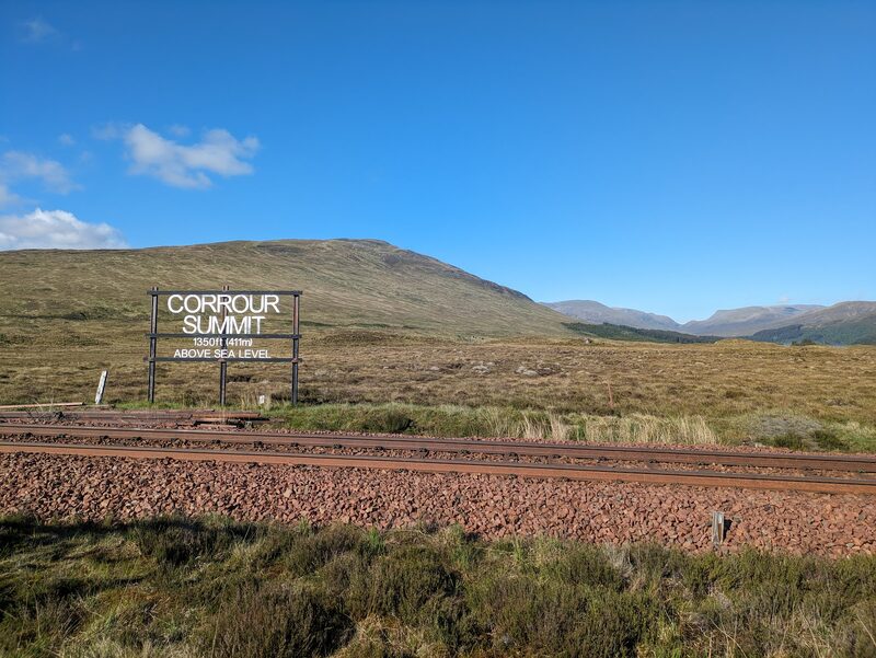 The Corrour Summit sign beside the railway line, a broad rounded hill behind it under a blue sky