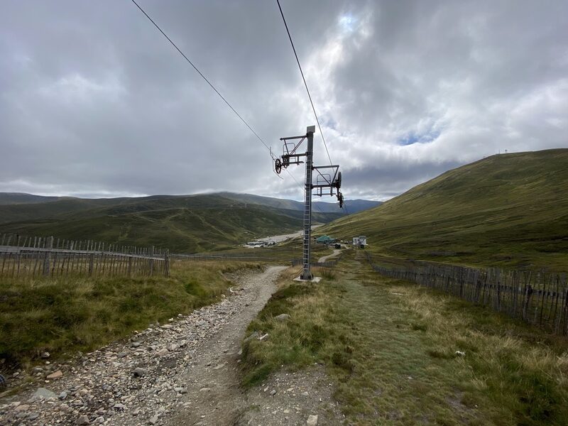A stony track between snow fences passing under a ski tow's pylon, the ski centre's buildings below and grey cloud overhead.