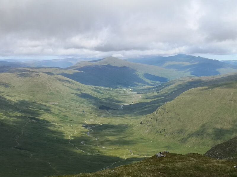 From high on Ben Challum, a long green glen winding away below under grey cloud, with hills in sun beyond.