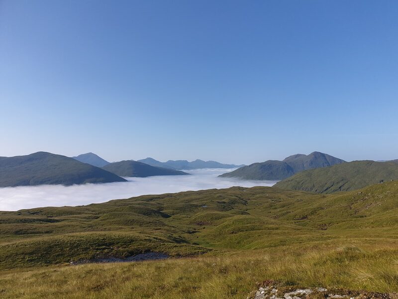A grassy shoulder with a small pool, and beyond it a sea of cloud with dark hilltops standing clear of it.