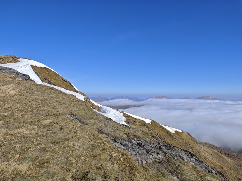The grassy upper slope of Fionn Bheinn with a band of old snow along the corrie rim, above a sea of cloud.