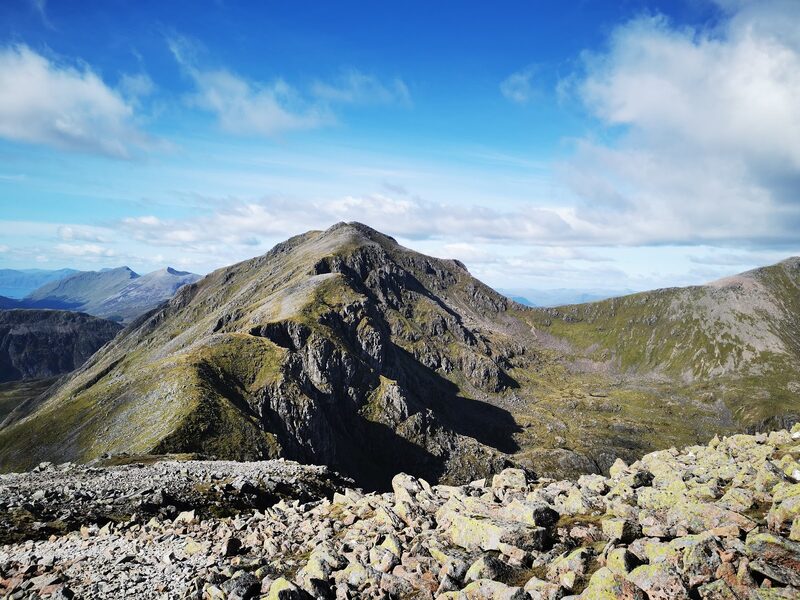 A rocky summit above dark crags on the Bidean nam Bian massif, seen across a field of boulders under a blue sky.
