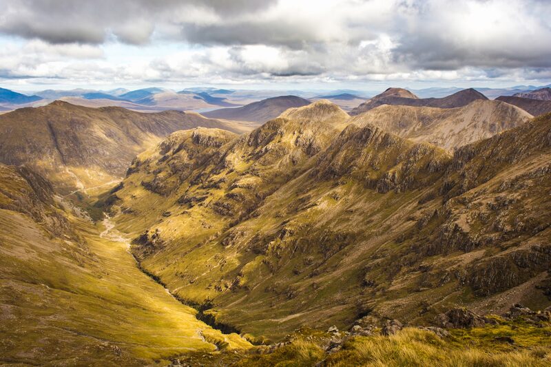 Looking down a long green glen from high on the hill, with ridges and far hills under broken cloud.
