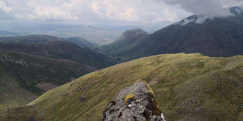 The view from a rock on the hill down a grassy shoulder to the glen and a big mountain in cloud.