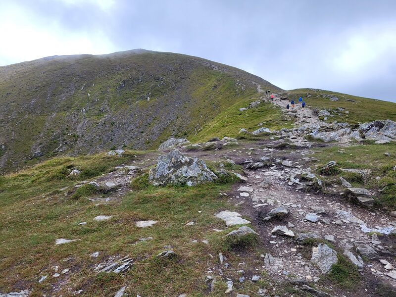 The stony path climbing towards the hill's upper slopes with cloud on the top, and a few walkers far up the path.