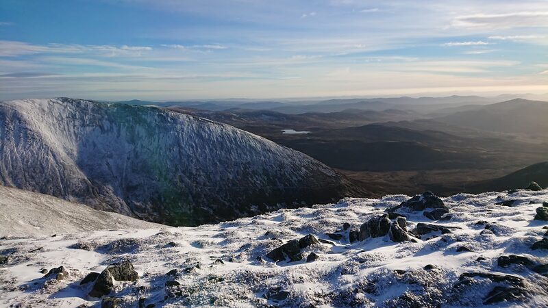 A rimed corrie wall falling from the summit, with the glens and low hills beyond in winter light.