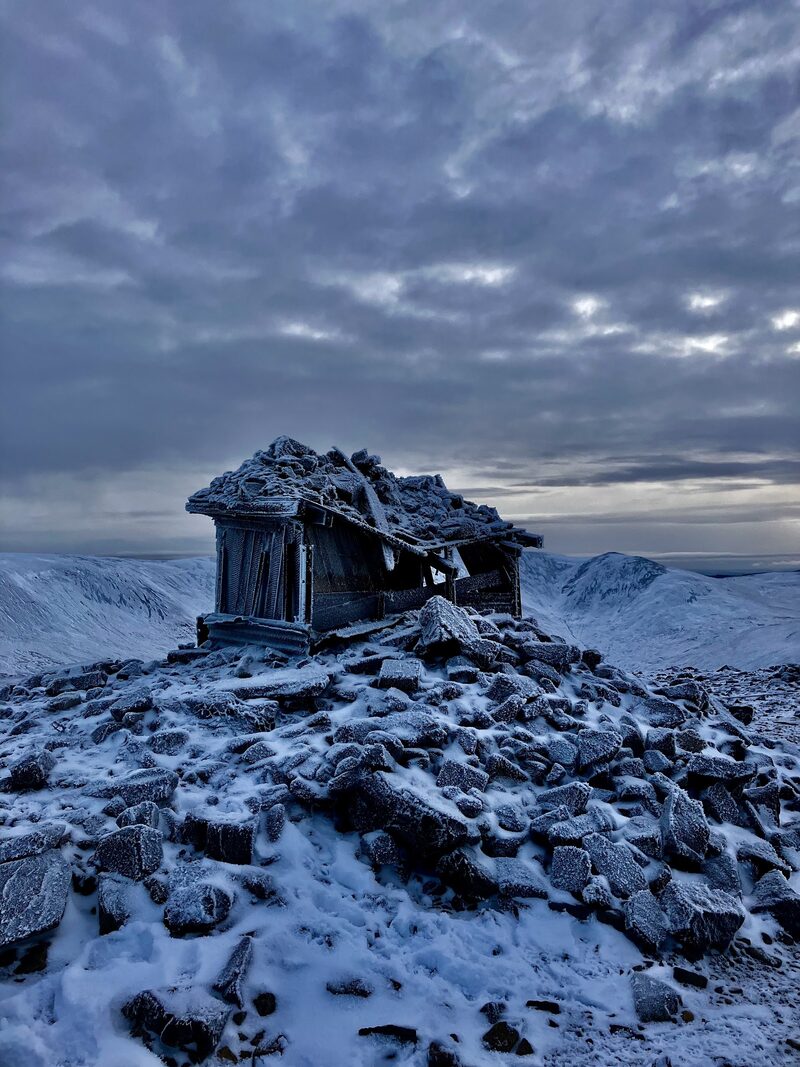 The small hut on the summit cairn of The Cairnwell thick with frost, snow on the stones and on the hills behind.