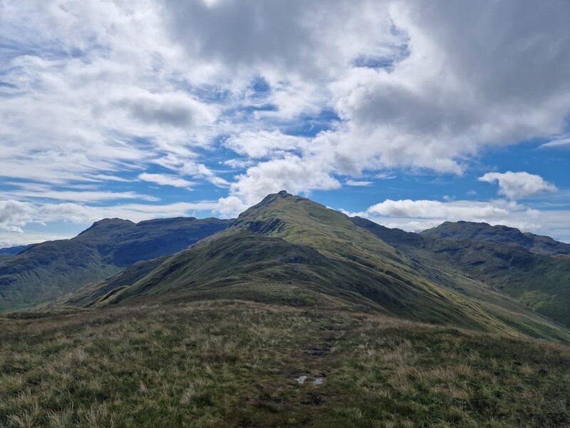A worn path along a grassy ridge to the pointed, rock-capped top of An Caisteal, with craggy hills either side under broken cloud.