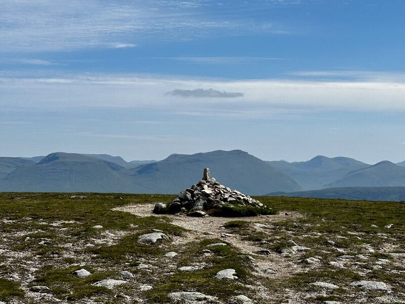 The summit cairn with a stone set on end on top, on a broad flat plateau of short grass and pale gravel, a line of blue hills along the skyline under thin cloud.