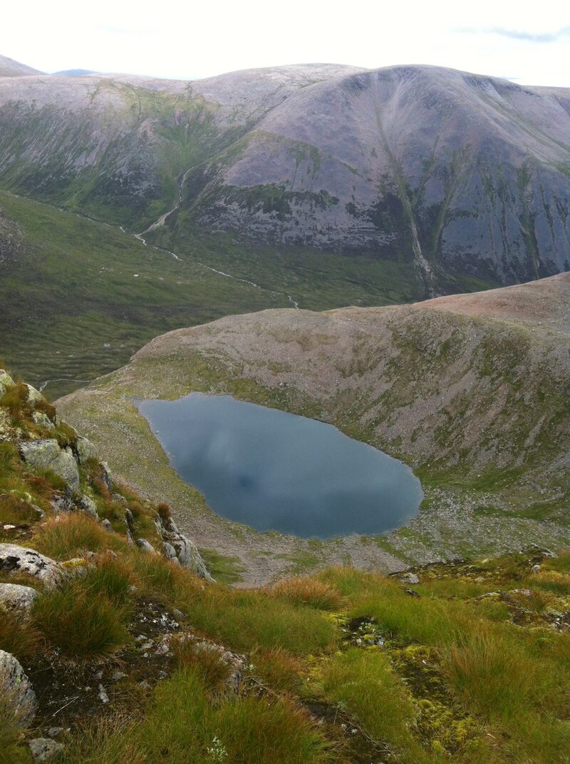 A dark lochan in its corrie seen from the grassy rocks above, with a deep glen and a slabbed hillside beyond.
