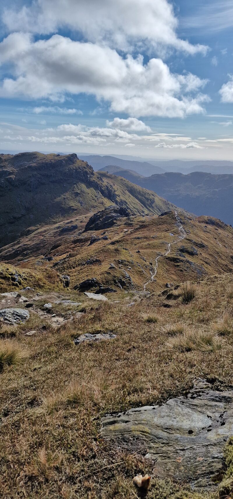 A path winding down a rocky ridge towards the long, crag-walled hill, under high white cloud.