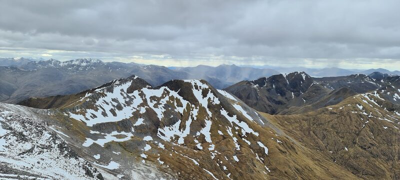 The view from high on the hill over snow-streaked ridges to far ranges under grey cloud.