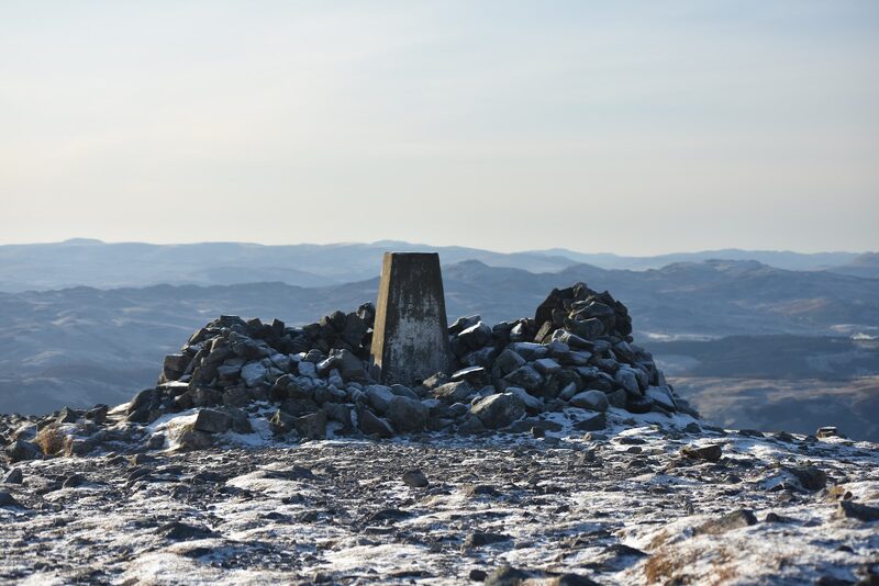 The trig pillar and cairn on the summit of Càrn Liath in frost, with blue hills beyond.