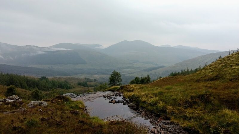 A pool of the burn on wet, tawny hillside, looking back down to the forest and the wide glen under cloud.