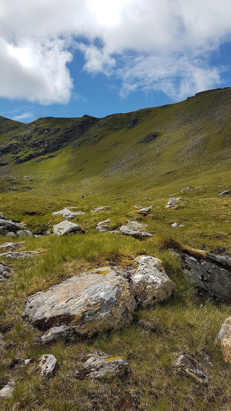 A boulder on the grassy slopes of A' Chailleach, the corrie wall and a blue sky behind.
